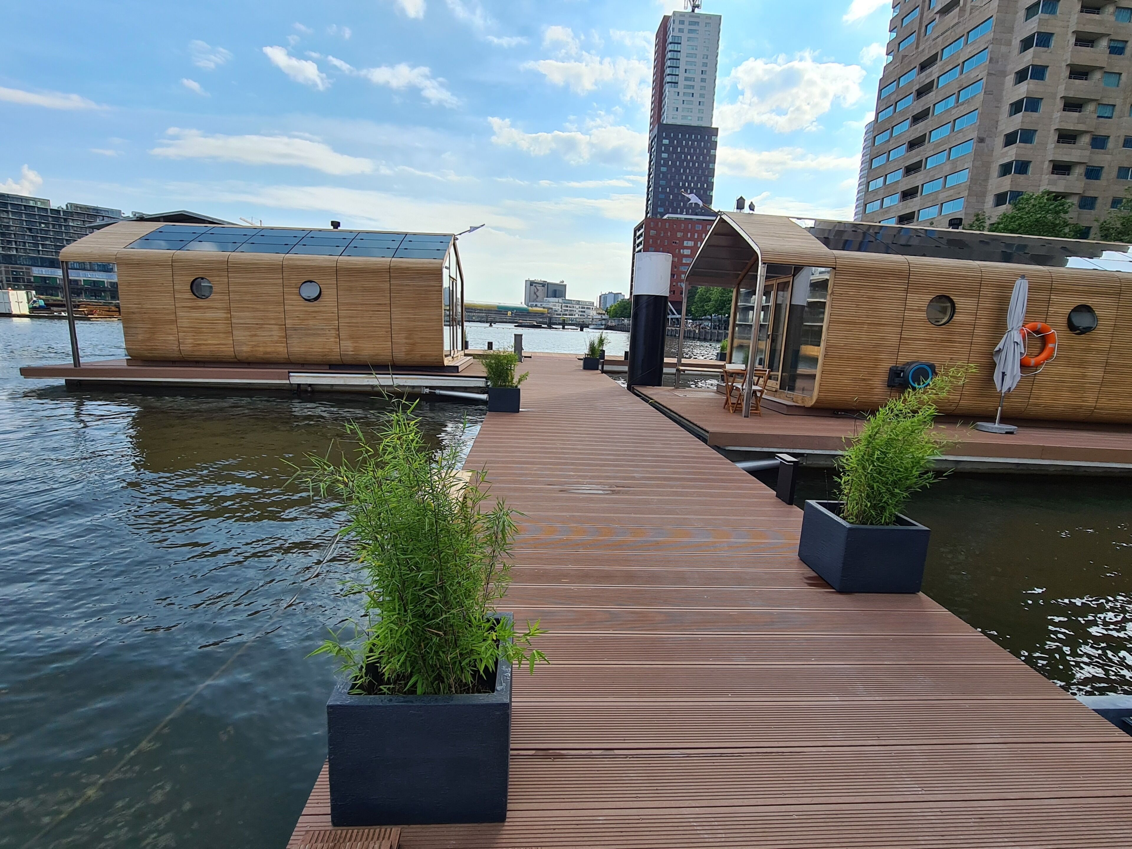 Una estructura flotante de madera con terraza, plantas en macetas y vistas al agua y al paisaje urbano.