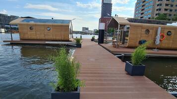 Una estructura flotante de madera con terraza, plantas en macetas y vistas al agua y al paisaje urbano.