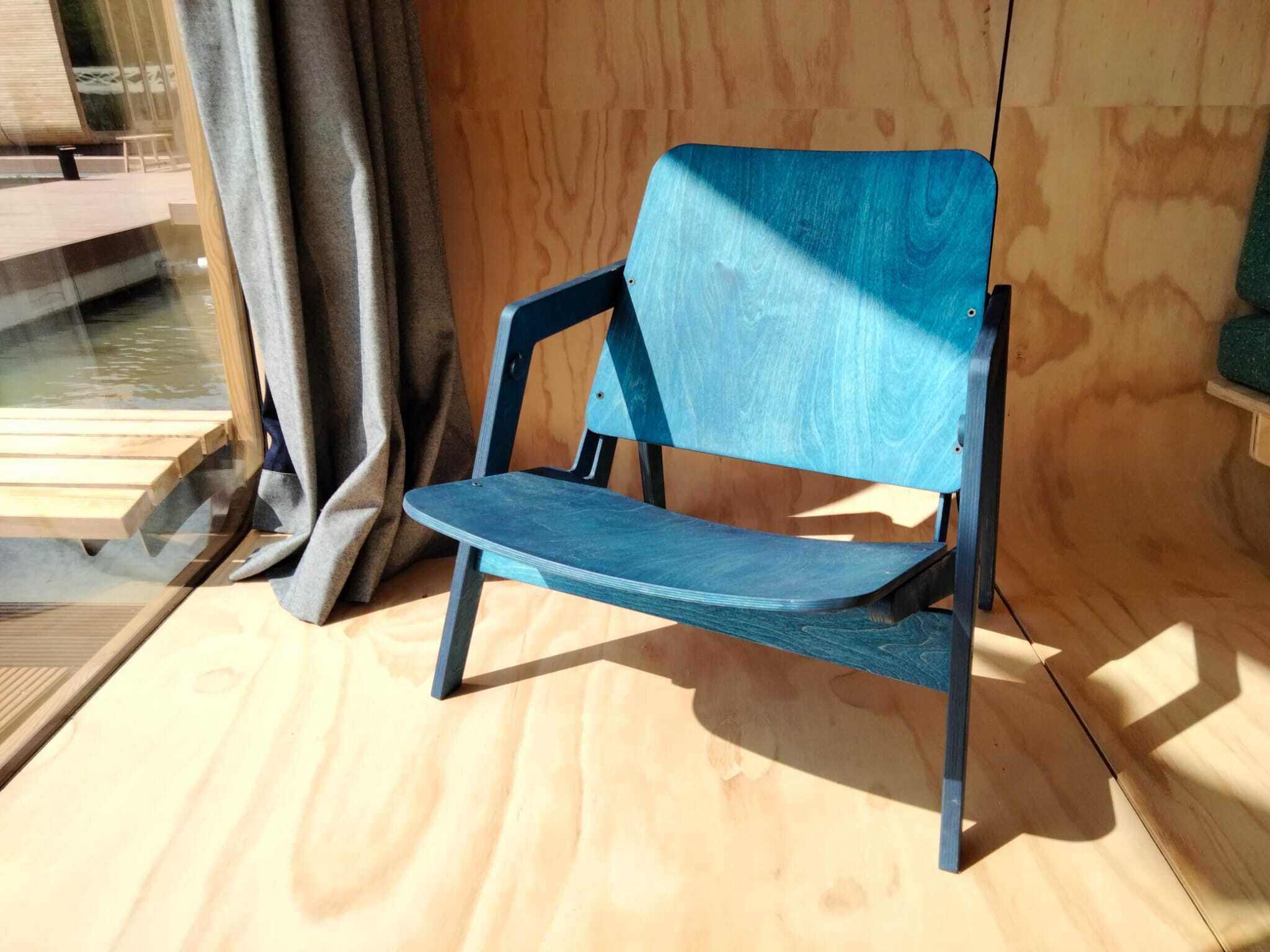 Ein moderner Stuhl mit Holzrahmen und blauer Sitzfläche, platziert in der Nähe eines Fensters mit Blick auf eine Holzterrasse.