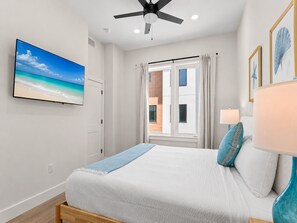 A bedroom with a bed, a ceiling fan, a window with curtains, a painting of a beach, and a lamp. - 3012 Emerald Coast A | Community Pool, Game Room + 0.3mi Walk to Beach (Panama City Beach)