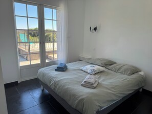 Ein Schlafzimmer mit Bett, Kissen, einem Nachttisch, einem Fenster mit Blick auf eine Terrass und einer Wandleuchte.