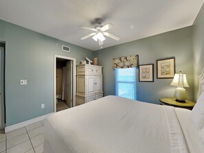 Une chambre à coucher comprenant un lit, un ventilateur de plafond, une fenêtre avec des stores, une commode et une table de chevet avec une lampe.