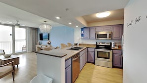 A modern kitchen with a central island, stainless steel appliances, and a dining area with wicker furniture. - Pelican Beach #2004 | Gulf Front in Destin + Amenities (Destin)