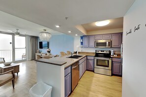 A modern kitchen with a central island, stainless steel appliances, and a dining area with wicker furniture. - Pelican Beach #2004 | Gulf Front in Destin + Amenities (Destin)
