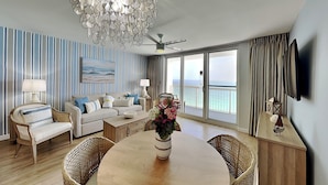A modern living room with a dining table, wicker chairs, a sofa, and a large window with a view of the sea. - Pelican Beach #2004 | Gulf Front in Destin + Amenities (Destin)
