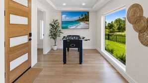 Um quarto com porta de madeira, mesa de pebolim, janela grande com vista para o verde e televisão na parede exibindo uma cena de praia.