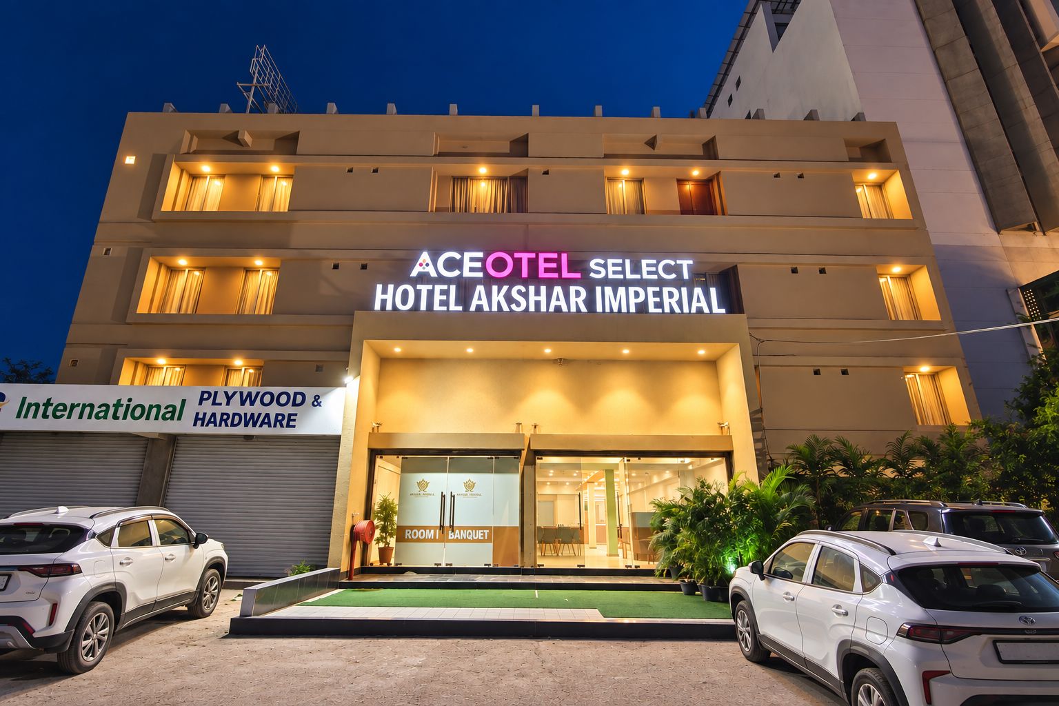 En hotelindgang med et skilt for 'ACEOTEL SELECT HOTEL AKSHAR IMPERIAL' og et skilt for 'International PLYWOOD & HARDWARE' i baggrunden.