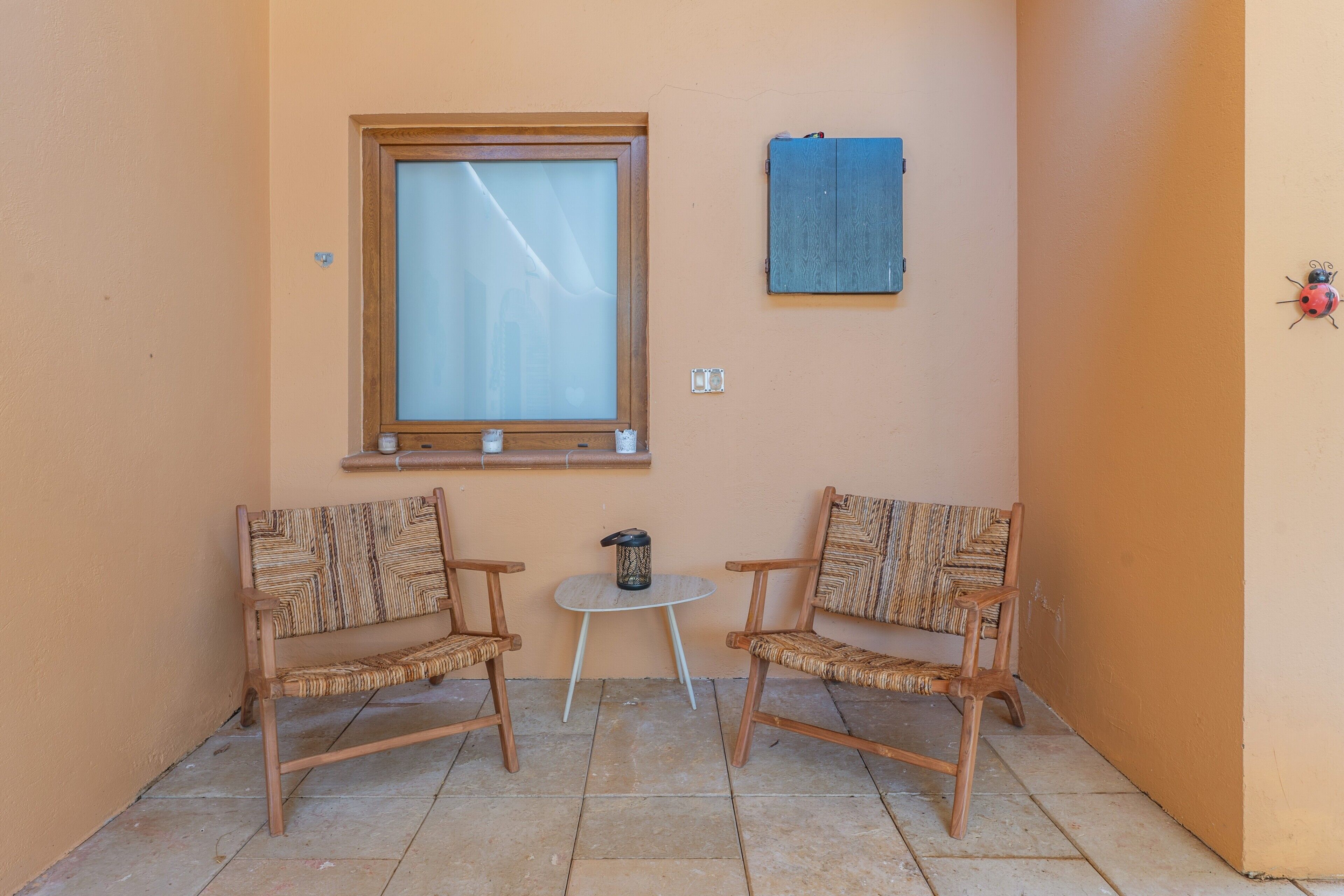 Una piccola stanza con due sedie in vimini, un tavolino rotondo, una finestra e un asciugamano appeso al muro.