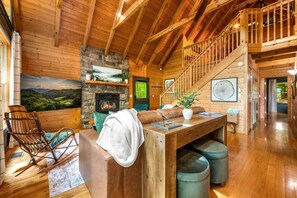 Ein gemütliches Blockhaus-Interieur mit Kamin aus Stein, Holzbalkendecke, einer Holztreppe und Blick ins Freie.