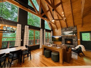 A cozy cabin interior with a wooden dining table, chairs, a fireplace, and large windows overlooking a forest. - 2BR w/Hot Tub*Fire Pit*Games*Privacy*Walk to Pool! (Sevierville)
