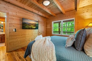 Ein Zimmer einer Holzjagdhuette mit einem Bett, einem Gemälde und einem Fenster mit Blick auf Grünfläche.