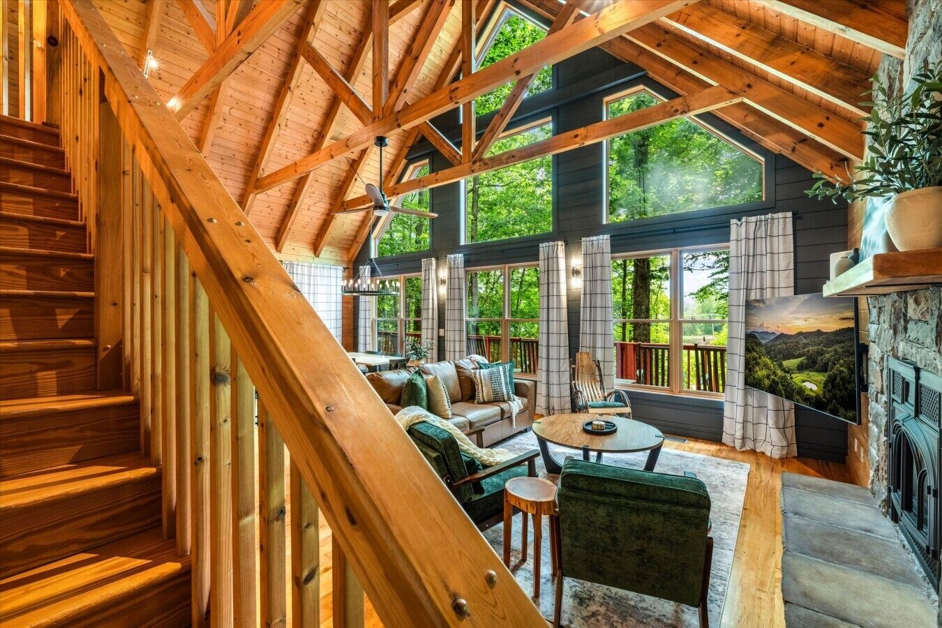 A spacious room with a high ceiling, wooden beams, a staircase, a sofa set, a fireplace, and large windows.
