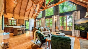 A spacious cabin interior with a wooden ceiling, large windows, a cozy seating area, and a kitchen. - 2BR w/Hot Tub*Fire Pit*Games*Privacy*Walk to Pool! (Sevierville)