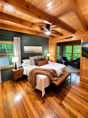 A wooden cabin bedroom with a bed, nightstands, a ceiling fan, and a view of the forest. - 2BR w/Hot Tub*Fire Pit*Games*Privacy*Walk to Pool! (Sevierville)