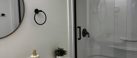 Un baño moderno con un lavamanos blanco, inodoro, ducha con mampara de vidrio y espejo con marco dorado.