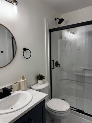 Et moderne badeværelse med en hvid vask, toilet, brusekabine i glas og et spejl med gylden ramme.
