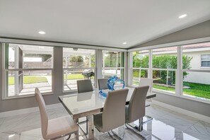 A modern dining area with a glass table and chairs, large windows, and a view of a garden.