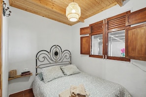 A bedroom with a bed, wooden ceiling, a hanging light fixture, a window with wooden shutters, and a wall-mounted shelf.