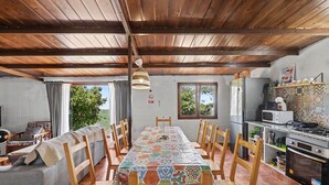 A dining area with a long table, wooden chairs, a patterned tablecloth, a hanging light fixture, a kitchen with appliances, and a view of greenery outside. - Finca El Lomo with Mountain Views, Private Pool and Wi-Fi (Saucillo)