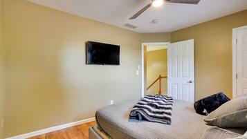 A bedroom with a bed, a television mounted on the wall, a ceiling fan, and a door leading to another room.