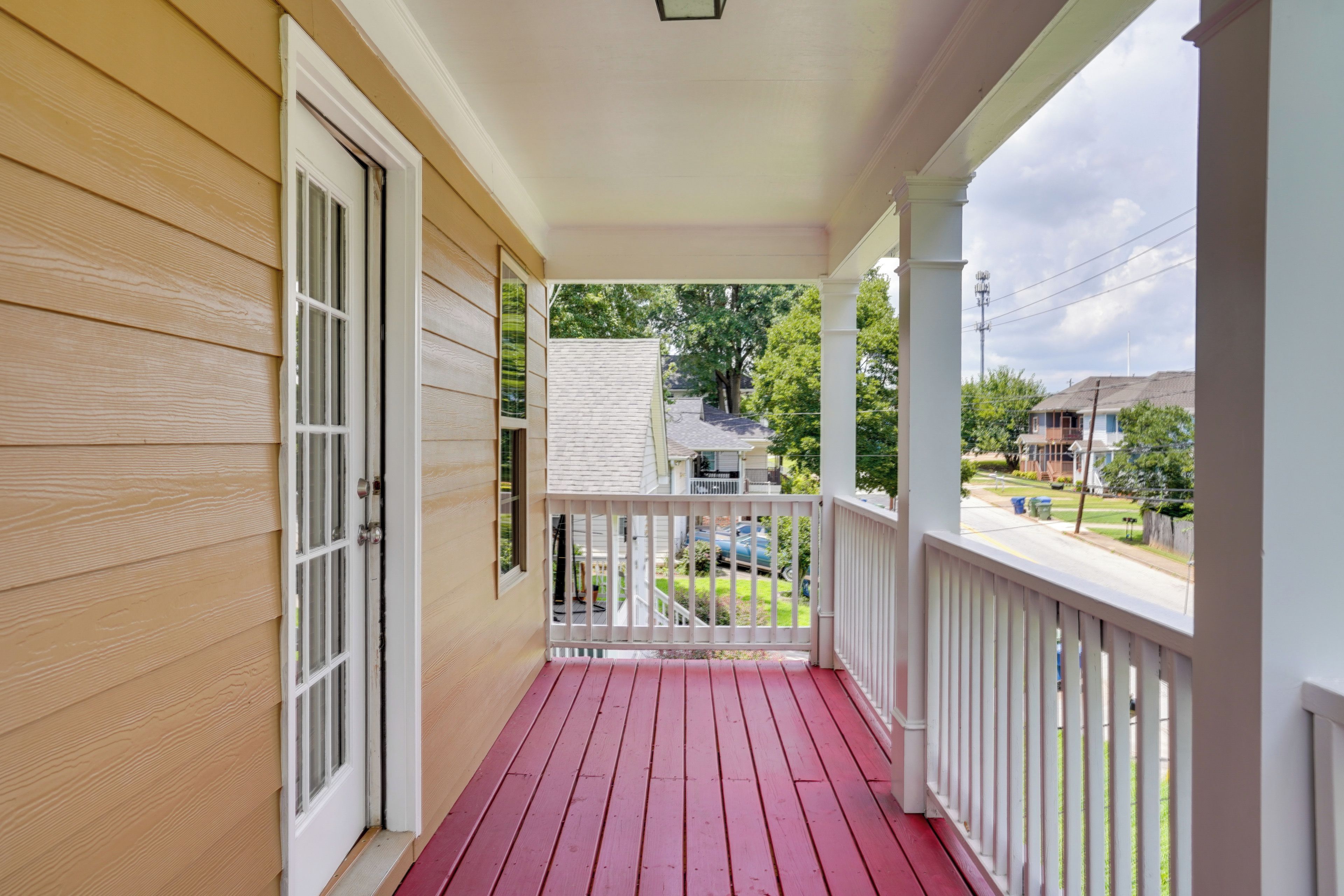 A wooden porch with a red floor, white railings, and a view of a residential street.