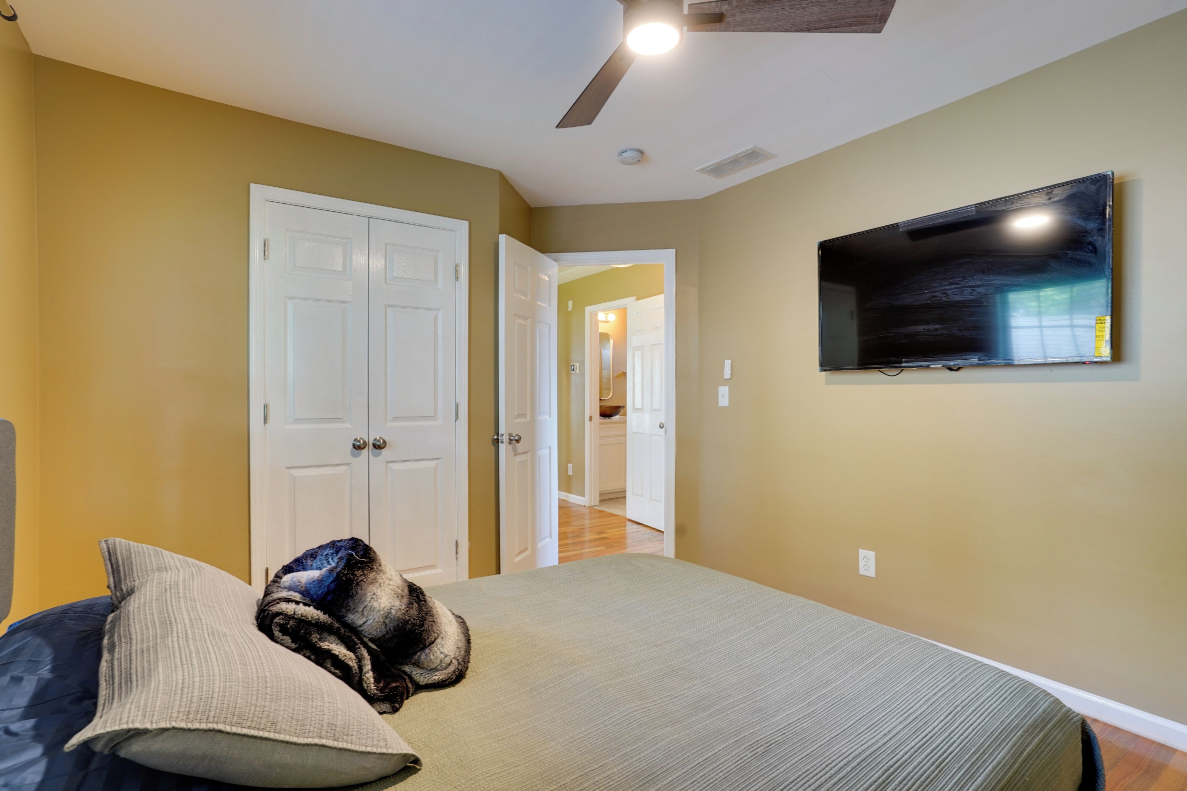 A bedroom with a bed, a television mounted on the wall, and a ceiling fan.