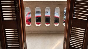 Blick durch eine offene Holztür mit Balkon, der zu einem weißen Gebäude mit schwarzen Fenstern und einem farbenfrohen Wandgemälde an der angrenzenden Mauer führt.