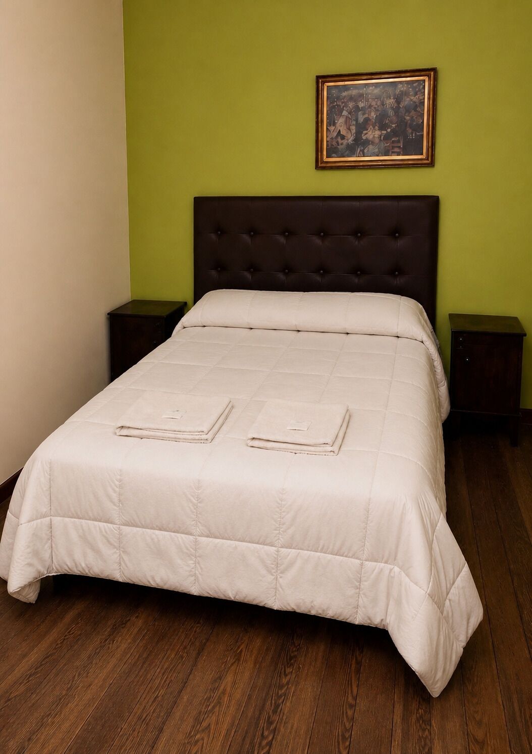 Una cama con edredón blanco acolchado, dos toallas dobladas, un cabecero tapizado oscuro, un cuadro en la pared y mesitas de noche de madera.