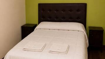 Una cama con edredón blanco acolchado, dos toallas dobladas, un cabecero tapizado oscuro, un cuadro en la pared y mesitas de noche de madera.