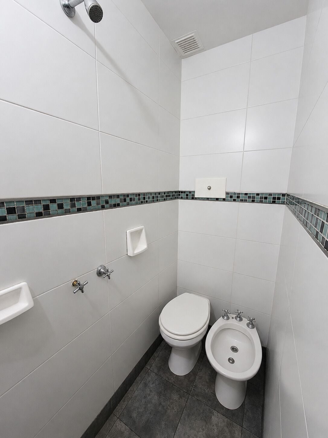 A small, modern bathroom with white tiles, a shower, a toilet, and a bidet.