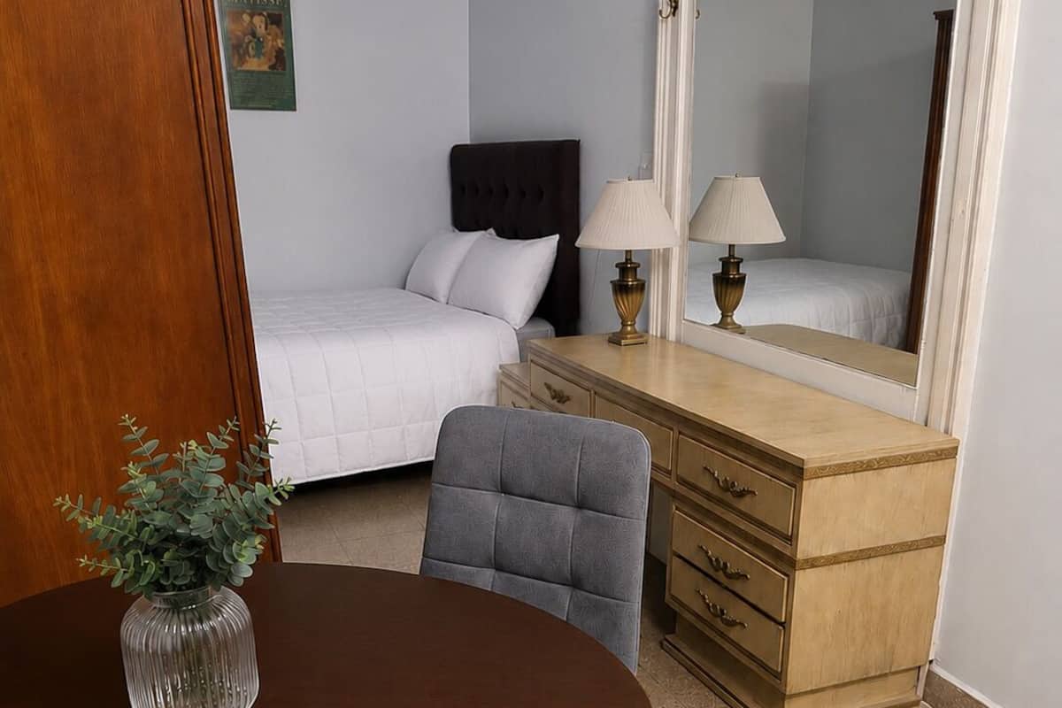A hotel room with a bed, a dresser, a mirror, a table with a vase, and a chair.