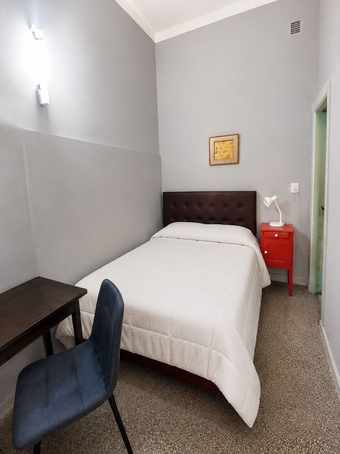 A bedroom with a bed, a desk, a chair, a bedside table with a lamp, and a framed picture on the wall.