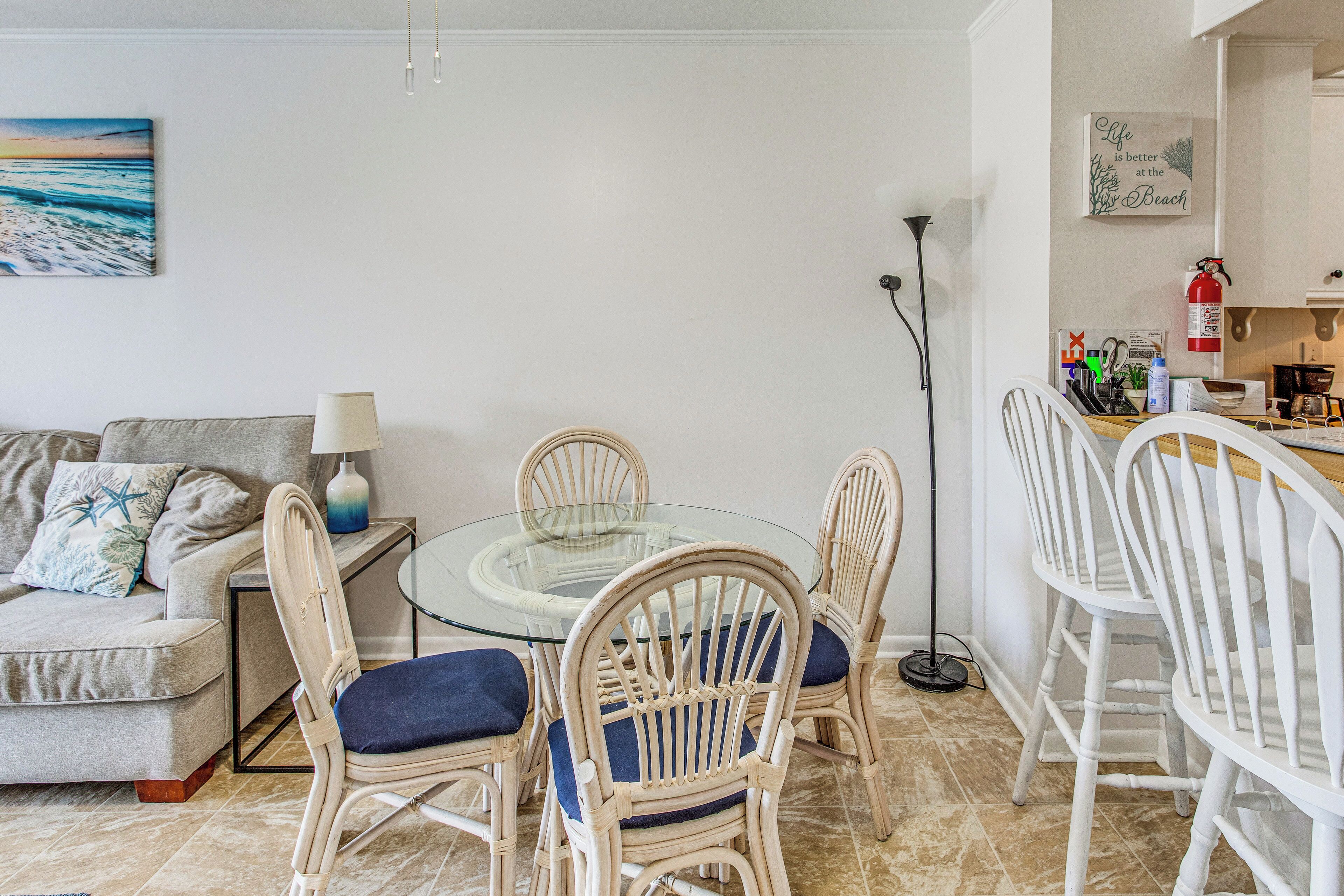 A dining area with a glass table surrounded by wicker chairs with blue cushions, a sofa with a lamp, and a wall with a painting and a sign.