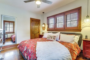 A bedroom with a large bed, wooden headboard, a ceiling fan, a mirror, and a window with blinds.