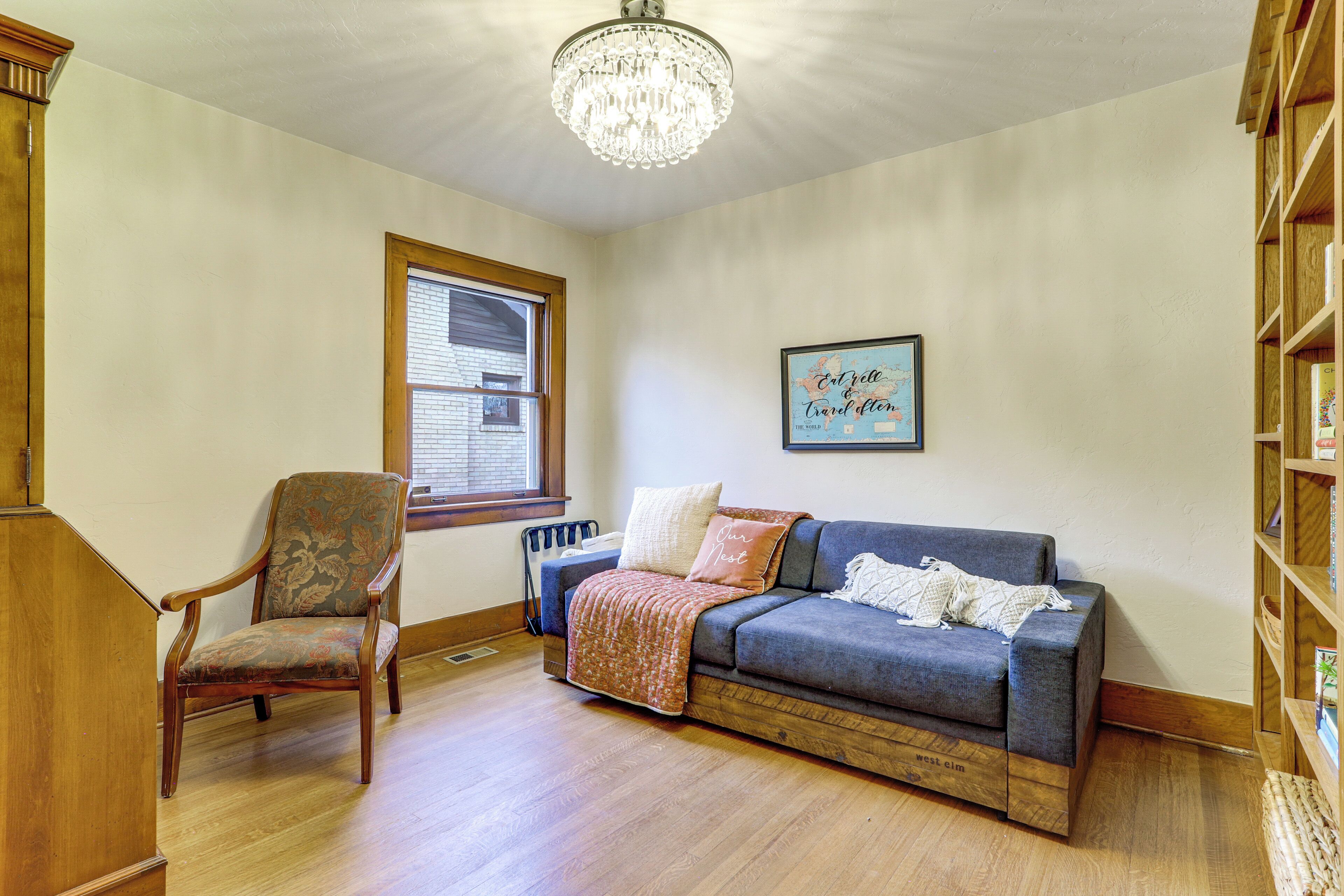 A room with a wooden floor, a blue sofa, a wooden chair, a chandelier, a window, and a bookshelf.