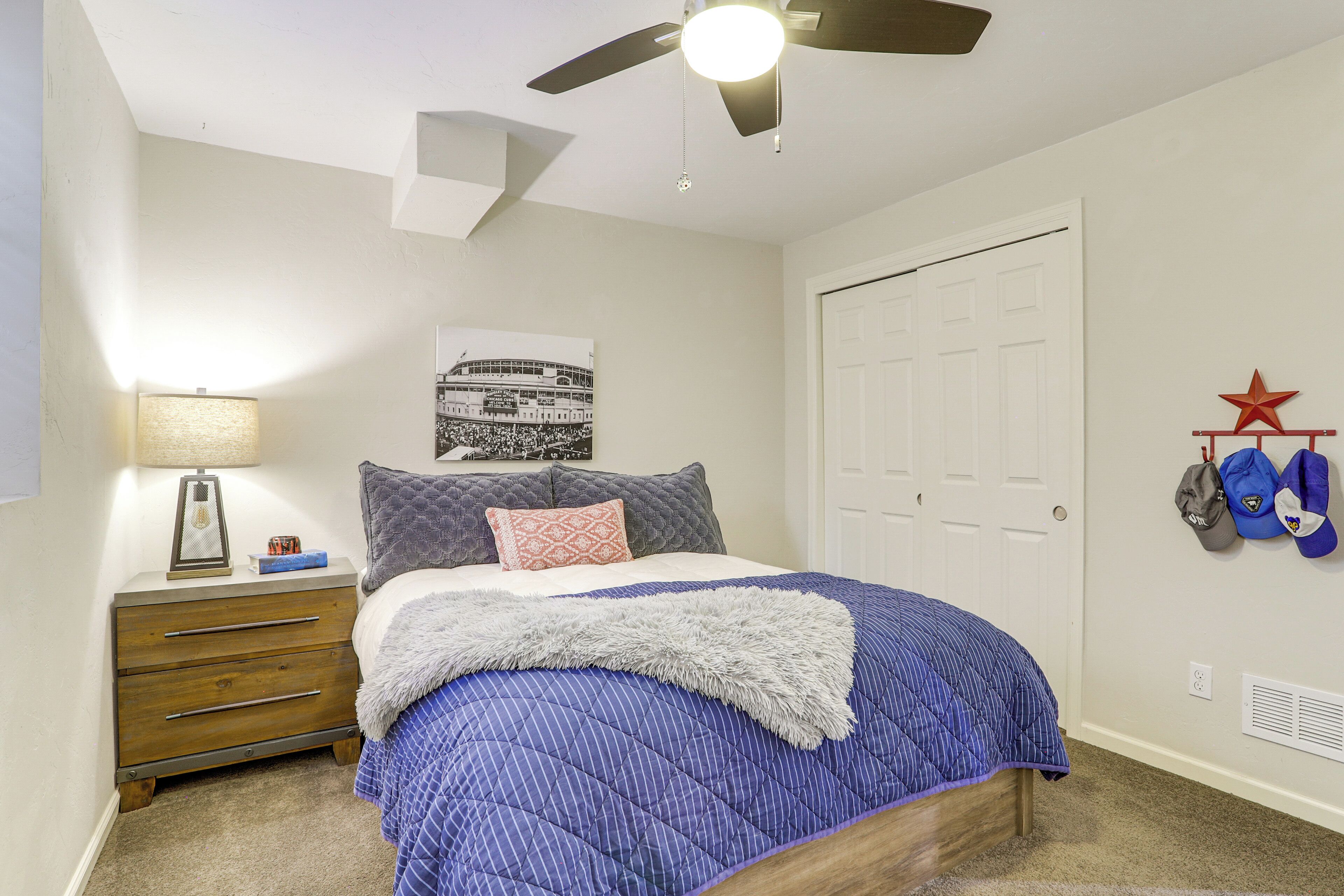 Un dormitorio con cama, mesita de noche, ventilador de techo, puerta y pared decorada con estrellas y tapas.