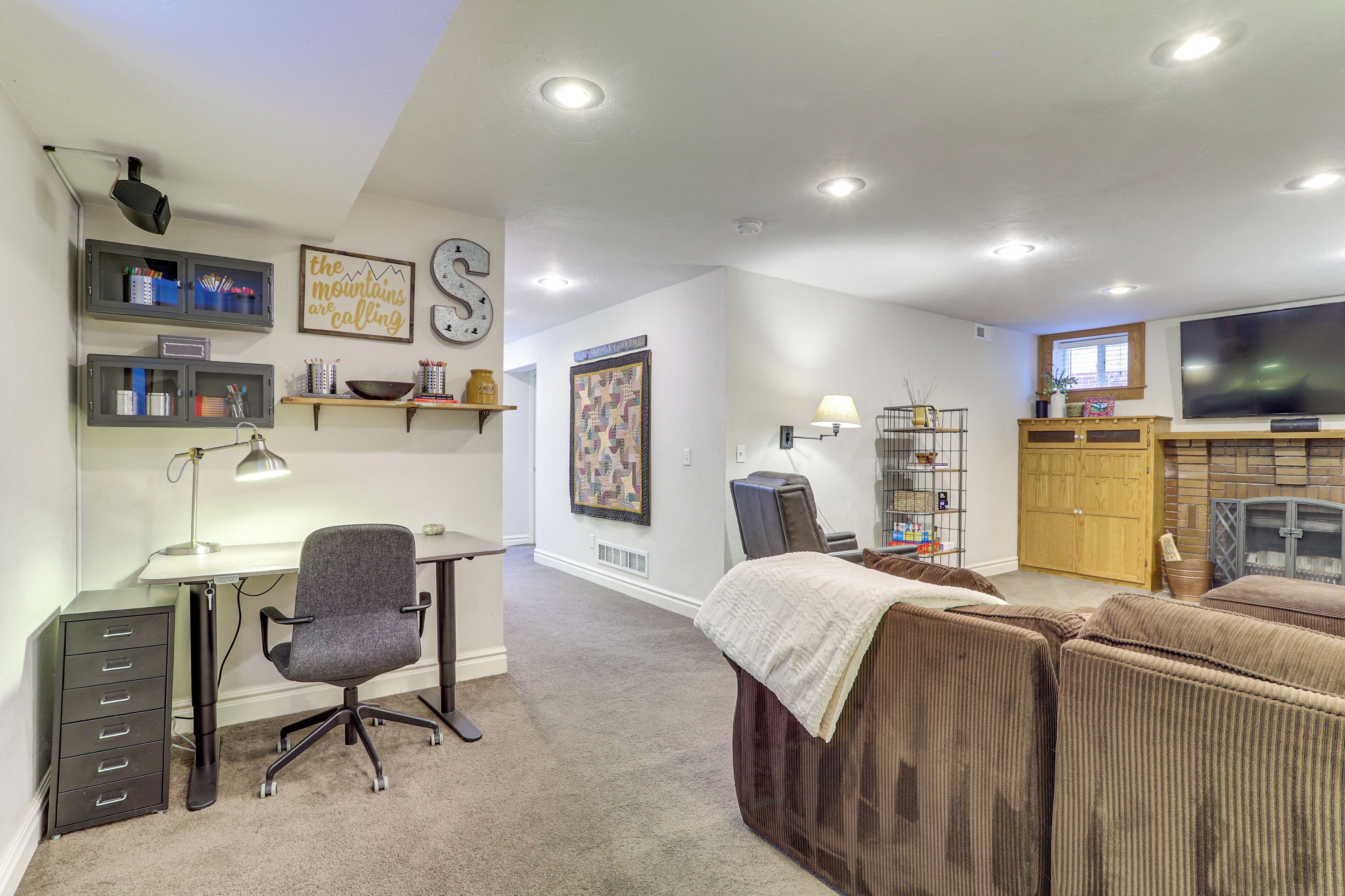 A well-lit room with a desk, office chair, and a large TV. There are shelves with various items and a fireplace.