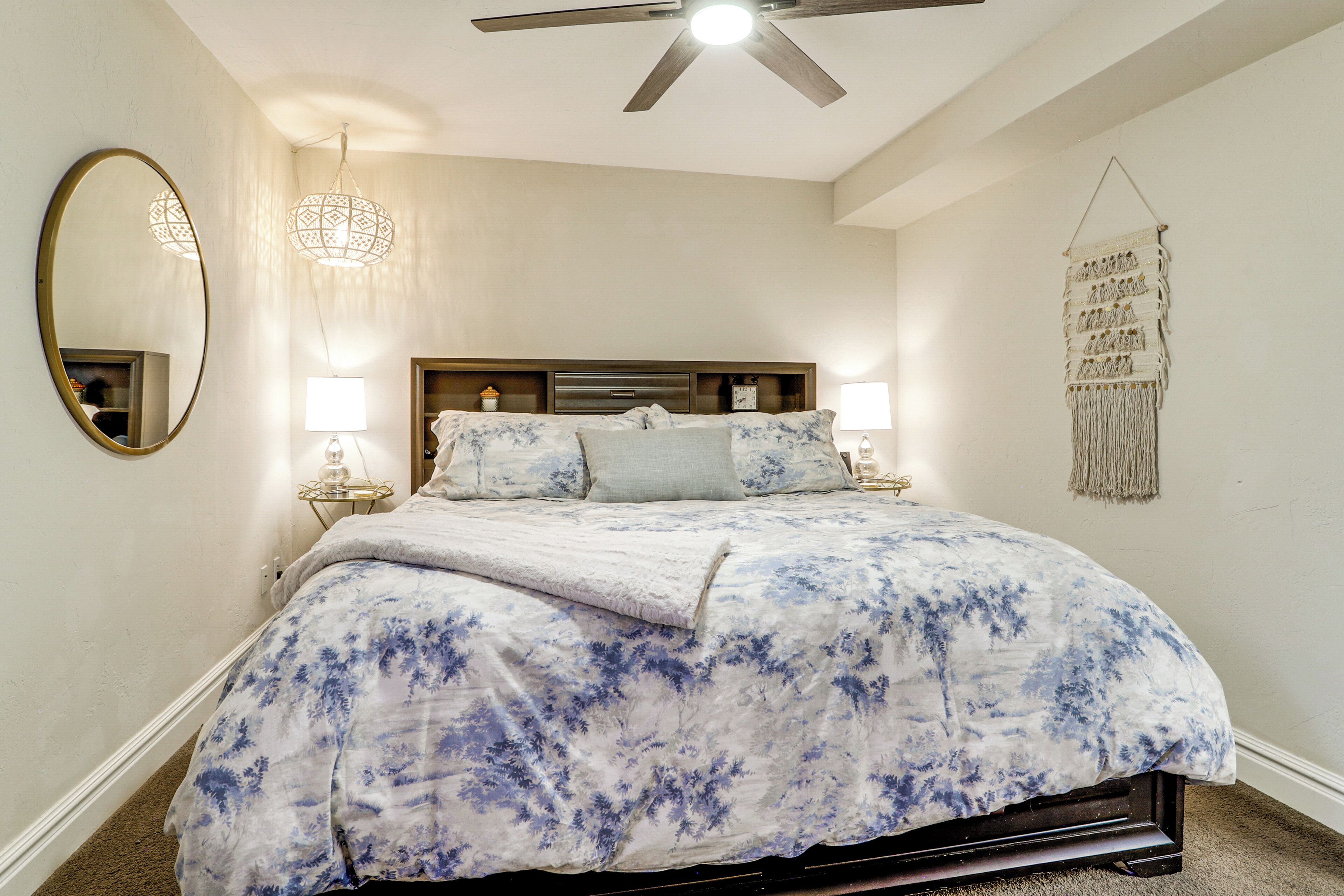 Une chambre à coucher comprenant un lit, un ventilateur de plafond, un miroir fixé au mur, des lampes de chevet et une décoration murale en macramé.