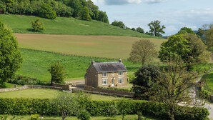 Ein Steinhaus mit Giebeldach, umgeben von einem eingezäunten Grundstück, eingebettet in eine grüne Landschaft mit Hügeln im Hintergrund.
