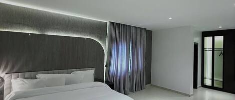 Un dormitorio moderno con una cama grande, una mesita de noche, una ventana con cortinas y una puerta que da a otra habitación.