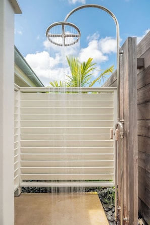 A modern outdoor shower with a curved metal frame, a white sliding door, and a wooden fence in the background. - Happy Shack - Heart of Hanalei (Hanalei)