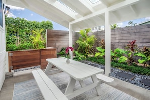 Un patio techado con una mesa y bancos de madera, rodeado por un jardín con diversas plantas y una valla de madera.