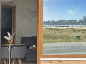 A room with a sofa, a small table with a vase, and a view of a beach with rocks and trees. - Flower Beach Helgueras 4 - 6094 (Noja)