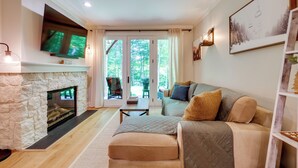 A living room with a stone fireplace, a flat-screen TV, a sofa with cushions, and a view through glass doors to an outdoor area. - Riverfront Ski-in/ski-out Condo at Loon Mountain (Lincoln)