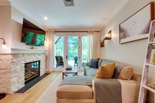 Una sala de estar con chimenea de piedra, un televisor de pantalla plana, un sofá con cojines y vistas a un espacio exterior a través de puertas de vidrio.
