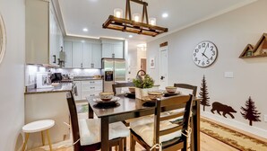 A modern kitchen with light blue cabinets, a stainless steel refrigerator, and a dining area with a wooden table and chairs. - Riverfront Ski-in/ski-out Condo at Loon Mountain (Lincoln)