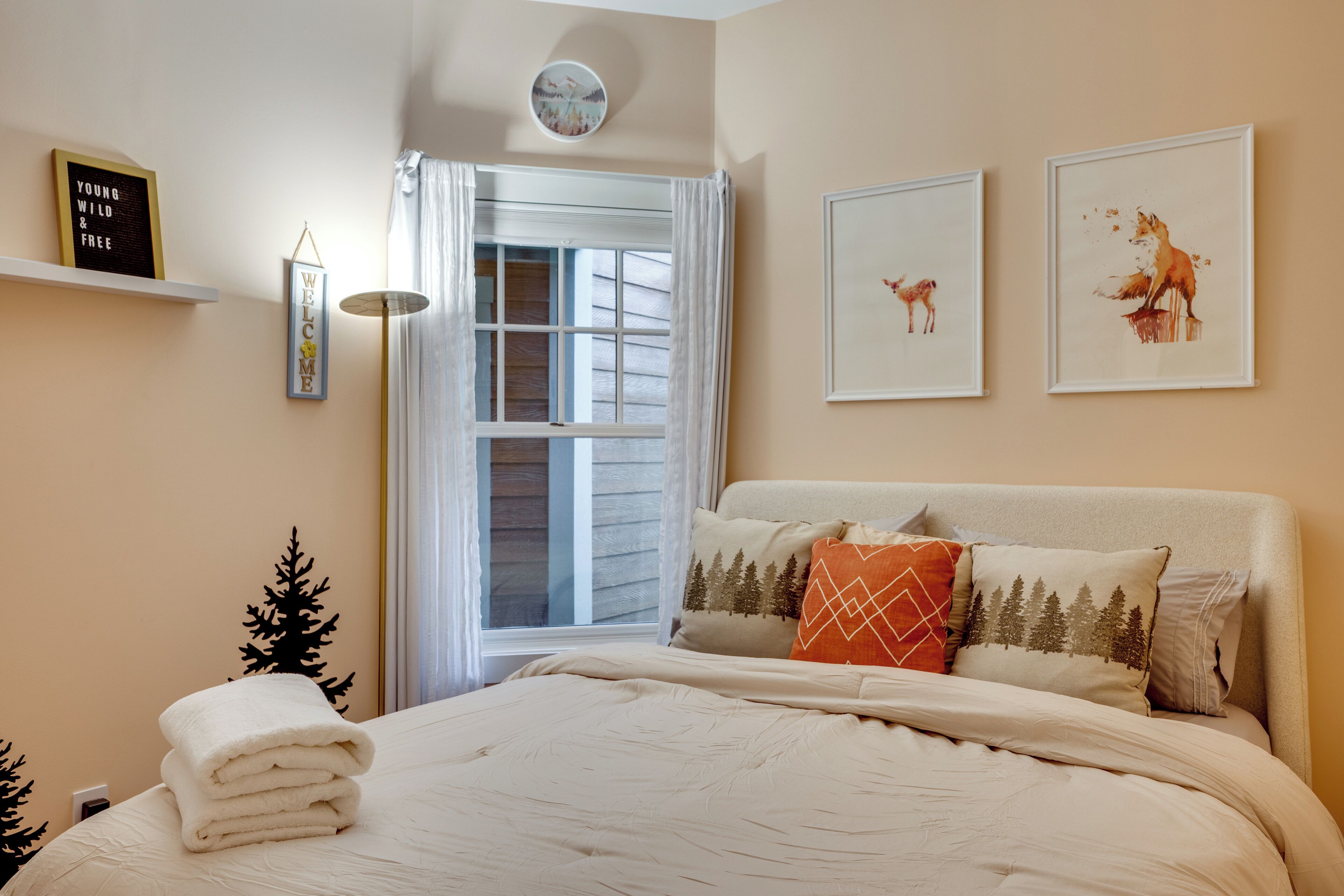 A bedroom with a bed, a window with curtains, a wall-mounted shelf with a sign, and framed pictures.