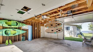 Un garaje abierto con kayaks y tablas de remo, suelo de hormigón y vista a una casa y un jardín.