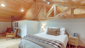 Un dormitorio con techo de madera, una cama con manta estampada, una lámpara de noche y una silla.