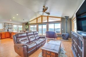 A spacious living room with a leather sofa, a wooden coffee table, a fireplace, and large windows offering a view of the outdoors. - South-facing Herron Island Retreat w/ Sound Views! (Lakebay)